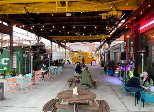 Outdoor dining area with colorful chairs, string lights, and food trucks, featuring people enjoying their meals.