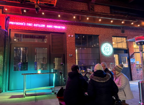 A group of people sitting outside a distillery with colorful lights and a sign reading "Providence's first distillery since Prohibition."