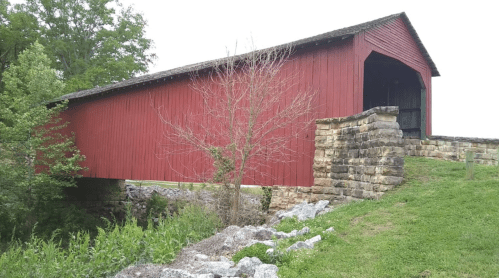 A red covered bridge stands over a small stream, surrounded by greenery and stone supports.