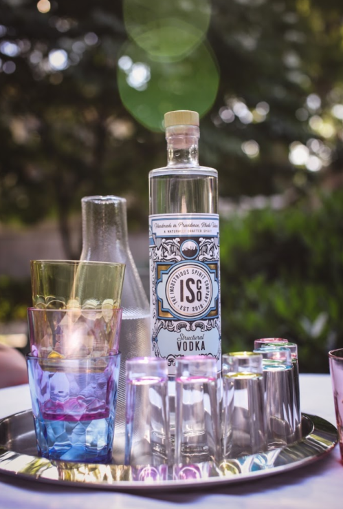 A bottle of vodka surrounded by colorful glasses on a tray, with a blurred outdoor background.