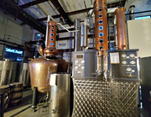 A copper and stainless steel distillation setup in a brewery, featuring large tanks and intricate piping.