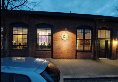 Exterior of a brick building with large windows, dimly lit, showing people inside. A sign reads "IS."