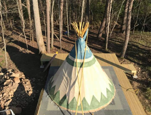 Aerial view of a colorful teepee on a wooden platform, surrounded by tall trees in a forested area.