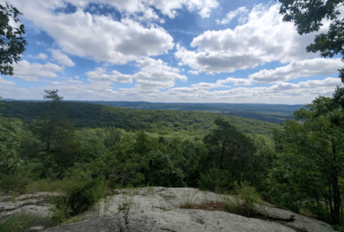 A panoramic view of lush green hills under a partly cloudy sky from a rocky overlook.