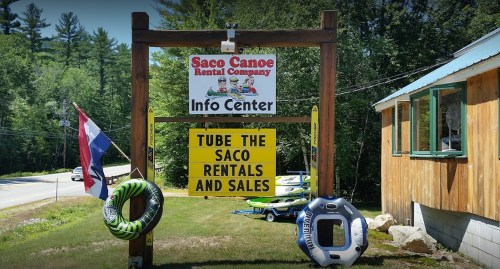Sign for Saco Canoe Rental Company with information about tubing, rentals, and sales, surrounded by greenery.