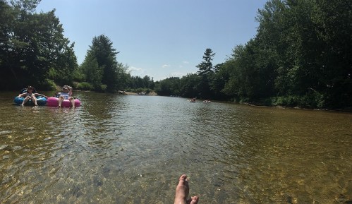 A serene river scene with people floating on tubes, surrounded by lush greenery and a clear blue sky.