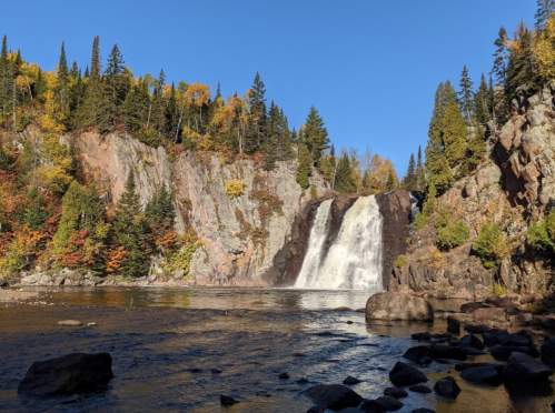 A scenic waterfall cascades down rocky cliffs, surrounded by vibrant autumn foliage and a clear blue sky.