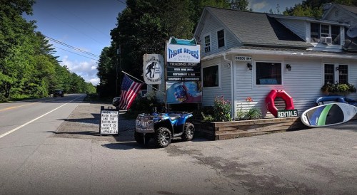 A roadside trading post with an American flag, ATV, and rental signs, surrounded by trees and a clear blue sky.