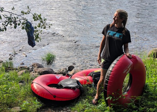 A woman stands by a river, holding a red inner tube, with another tube and a shirt hanging from a tree nearby.
