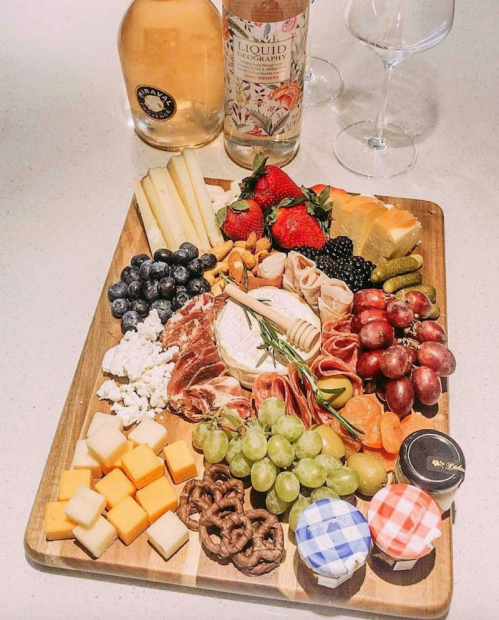 A wooden platter filled with assorted cheeses, fruits, nuts, meats, and pretzels, accompanied by wine bottles and glasses.