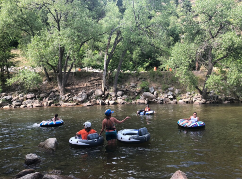 A group of people floating on inner tubes in a river, surrounded by trees and rocky banks on a sunny day.