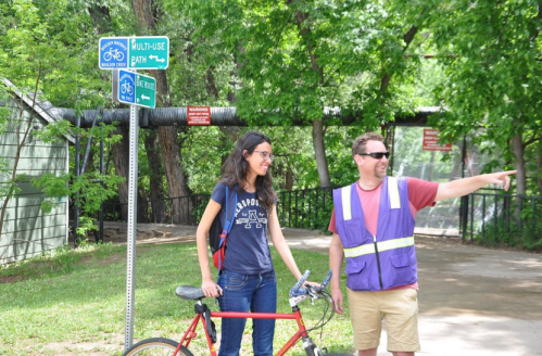 A man in a vest points while a woman with a bike smiles, near signs for a multi-use path in a green park.