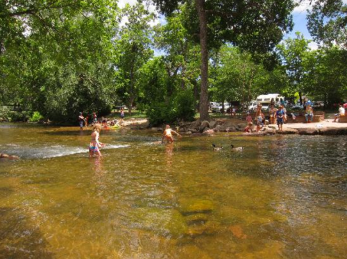 A sunny riverside scene with people swimming, playing, and relaxing among trees and a clear stream.