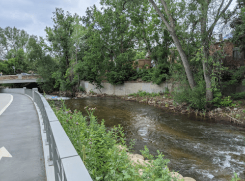 A serene river flows beside a pathway, surrounded by lush greenery and trees under a cloudy sky.