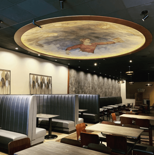 Interior of a modern restaurant featuring booth seating, wooden tables, and a decorative ceiling mural.