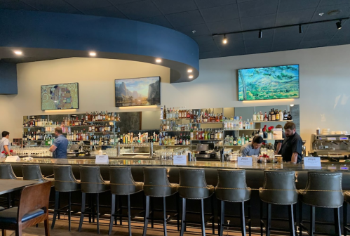 A modern bar with a long counter, shelves of drinks, and two large screens displaying artwork. Staff are preparing drinks.