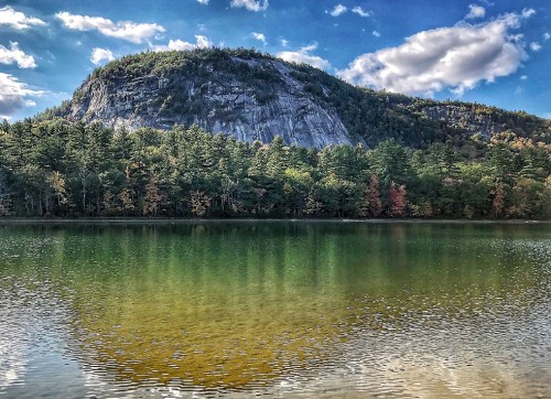 A serene lake reflects a rocky mountain and colorful trees under a blue sky with fluffy clouds.