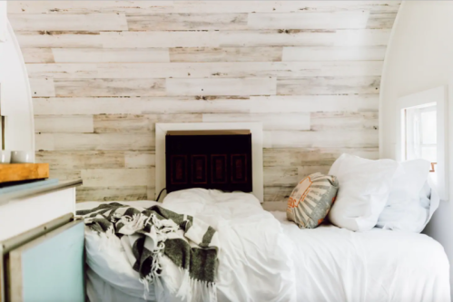 Cozy bedroom with a white bed, textured wood wall, and a decorative pillow, featuring soft blankets and natural light.
