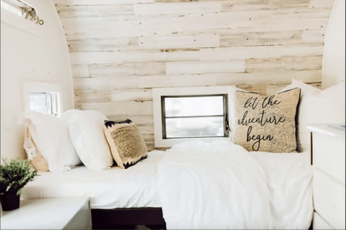 Cozy bedroom with white bedding, decorative pillows, and wooden walls, featuring a motivational throw pillow.
