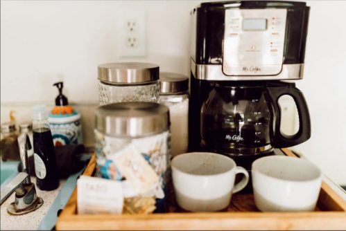 A coffee maker on a wooden tray with jars, sugar, and two white cups beside it.