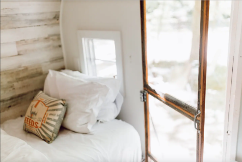 Cozy bedroom with a white bed, decorative pillow, and a wooden door leading to an outdoor view.