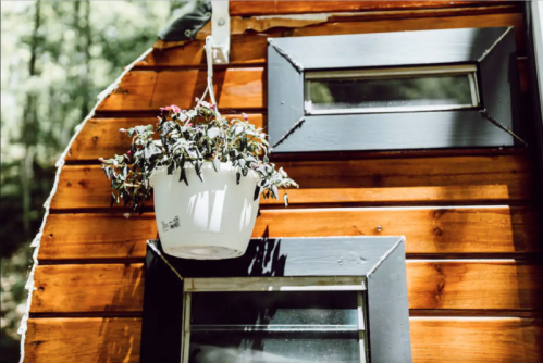 A hanging white planter with plants is attached to a wooden wall with a small window. Sunlight casts shadows.