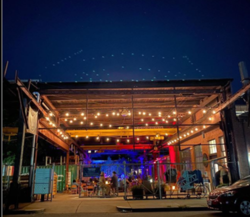 A lively outdoor venue at night, illuminated by string lights and colorful lights, with people enjoying the atmosphere.