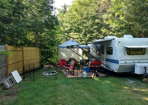 A cozy campsite with a trailer, chairs, and a canopy surrounded by trees and a wooden fence.