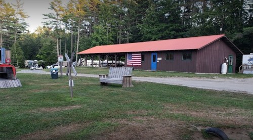A rustic building with a red roof and American flag, surrounded by trees and a grassy area with benches.