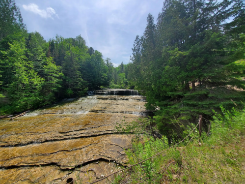 A serene landscape featuring a flowing river over rocky terrain, surrounded by lush green trees under a clear blue sky.