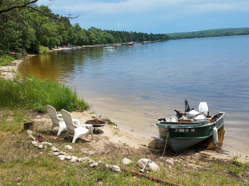 A peaceful lakeside scene with a small boat, two chairs, and a fire pit on the sandy shore surrounded by greenery.