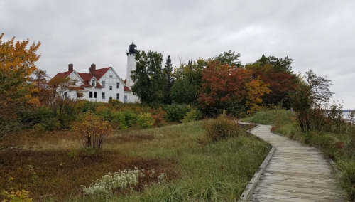 A wooden path leads through colorful foliage to a white lighthouse and historic building under a cloudy sky.