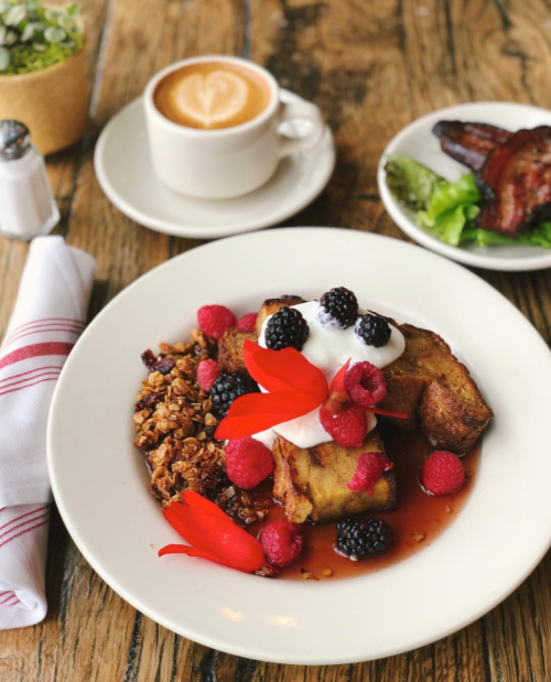 A plate of French toast topped with berries, yogurt, and edible flowers, alongside a cup of coffee and a side of bacon.
