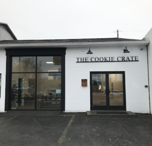 Exterior of a building with large glass doors and a sign that reads "The Cookie Crate." Parking lot in front.