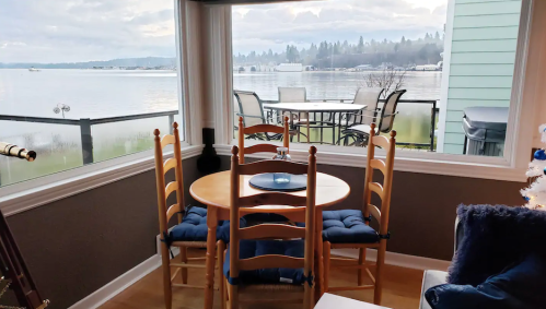 Cozy dining area with a round table and chairs, overlooking a serene lake view through large windows.