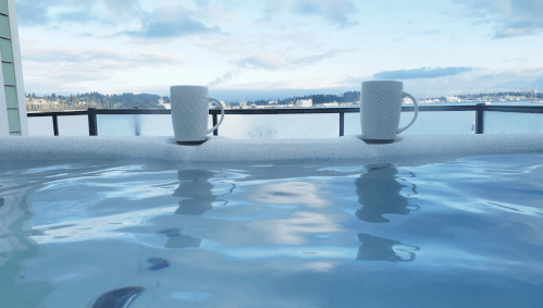 Two white mugs sit on the edge of a hot tub, reflecting in the water with a scenic view of the lake and sky.