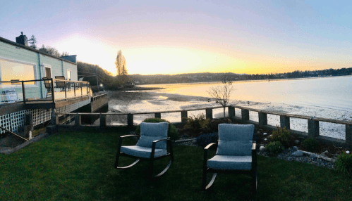 Two rocking chairs on a lawn overlooking a calm bay at sunset, with a house and trees in the background.