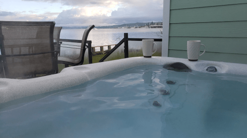A hot tub with clear water, two mugs on the edge, and a scenic view of the lake and mountains in the background.