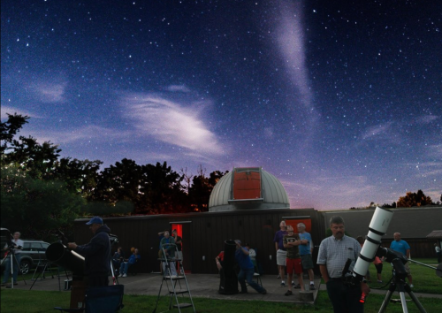 A group of people gather outside an observatory under a starry sky, using telescopes and enjoying the night.