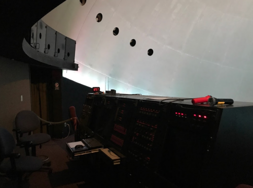 Control room with dark panels, buttons, and a flashlight, set against a curved wall. Two empty chairs are visible.