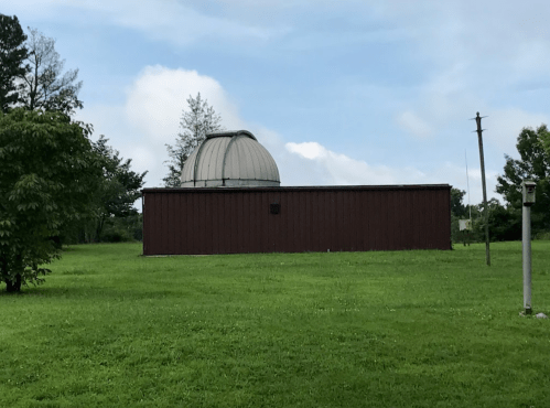 A brown observatory building with a dome, set in a grassy area under a cloudy sky.