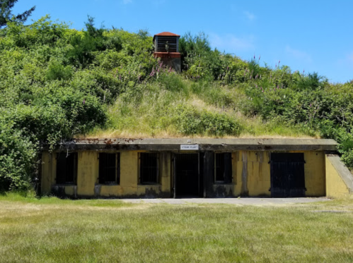 A weathered concrete bunker with barred windows, set against a grassy hill and blue sky. A lookout tower is visible above.