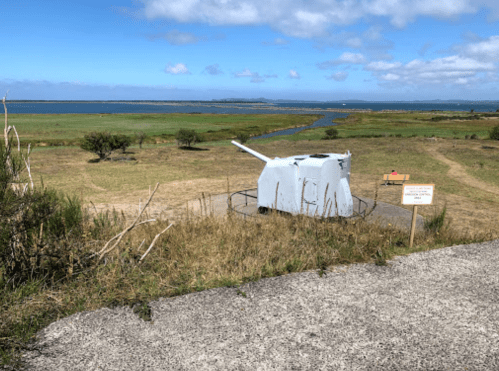 A coastal landscape featuring a white artillery gun on a grassy area, overlooking a river and the ocean under a blue sky.