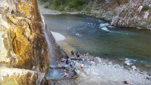 A natural hot spring by a river, with people relaxing in the water and enjoying the scenic surroundings.