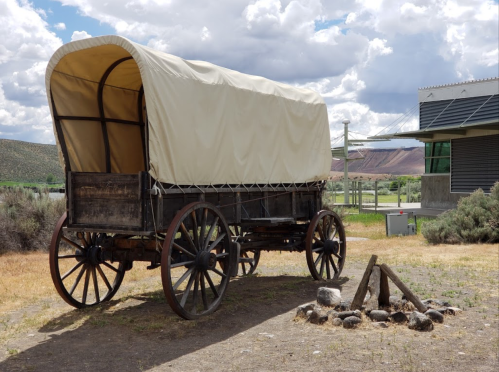 A covered wooden wagon stands beside a small campfire in a grassy area with clouds in the background.