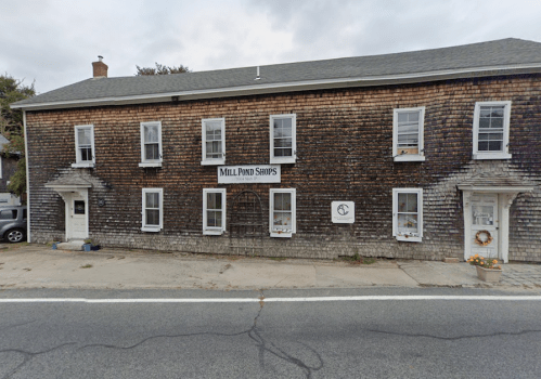 A rustic two-story building with wooden siding, featuring shops and a sign that reads "Mill Pond Shops."