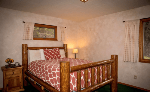 Cozy bedroom with a wooden bed, patterned bedding, and a lamp, featuring natural light from a window.