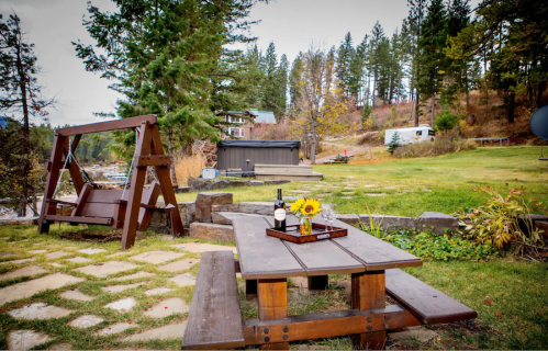 A wooden picnic table with a wine bottle and flowers, surrounded by trees and a scenic outdoor area.