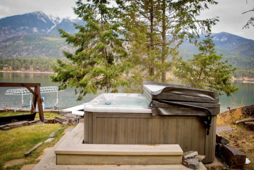 A hot tub on a wooden deck surrounded by trees, overlooking a lake and mountains in the background.