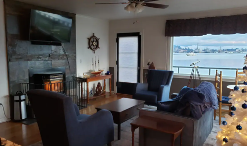 Cozy living room with a TV, fireplace, and a view of the water, featuring blue chairs and a decorated Christmas tree.